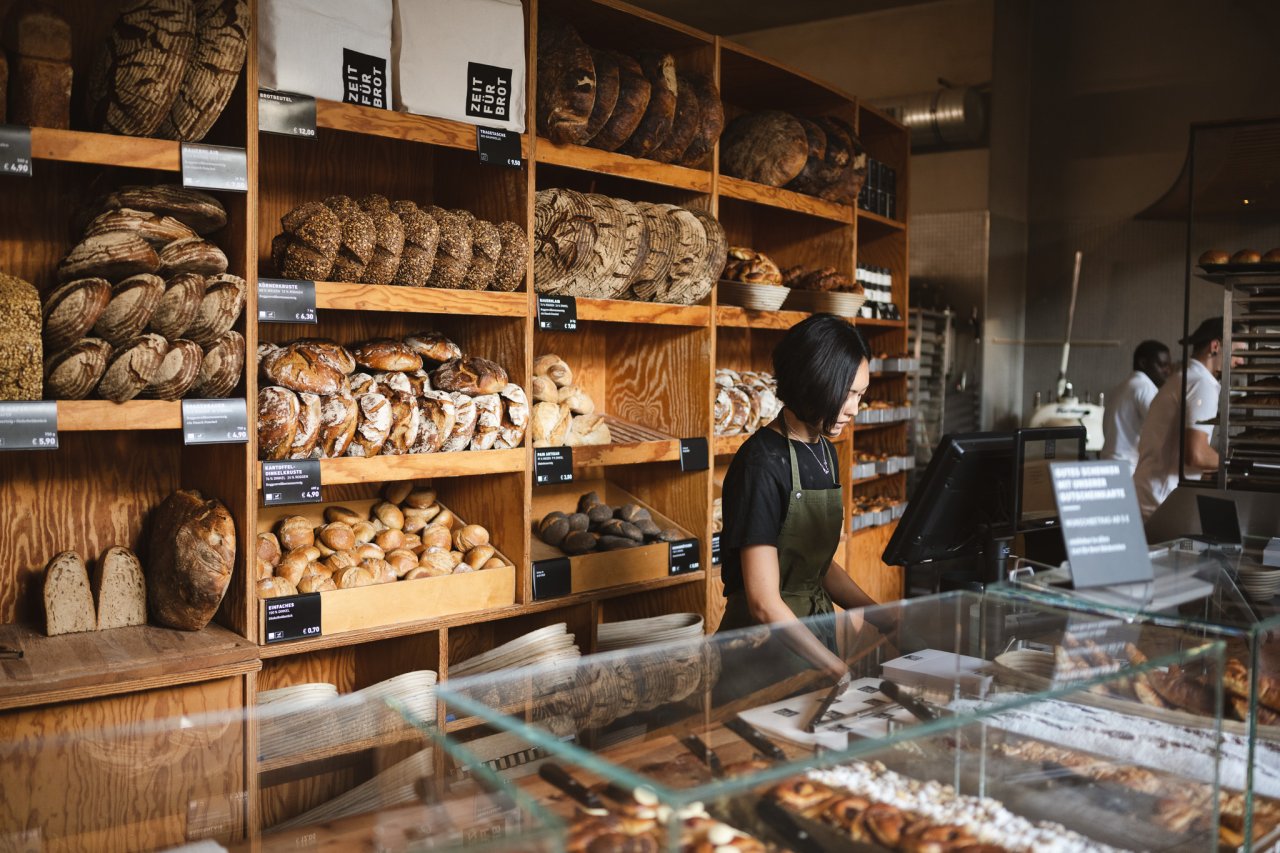 Holzregal mit verschiedenen Brotsorten und Br&ouml;tchen. Im Vordergrund eine Glasvitrine mit frischen Backwaren.